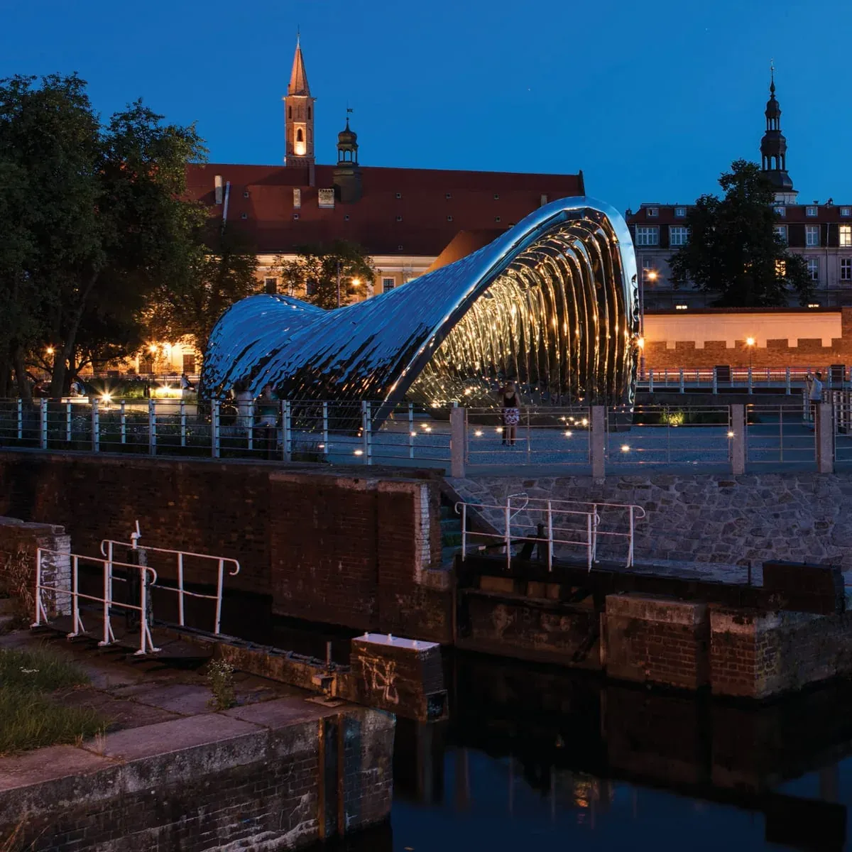 Sculpture of polished steel arches on a riverbank at dusk